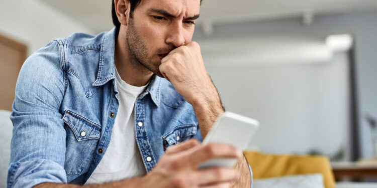 Young man feeling worried while reading text message on mobile phone at home.